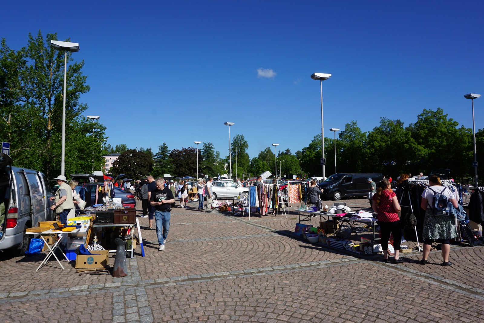 Kesäinen kirpputoripäivä Kouvolan Pohjola-torilla. Paljon myyjiä ja ihmisiä tutkimassa myytäviä tuotteita. Aurinko paistaa ja taivas on selkeä.