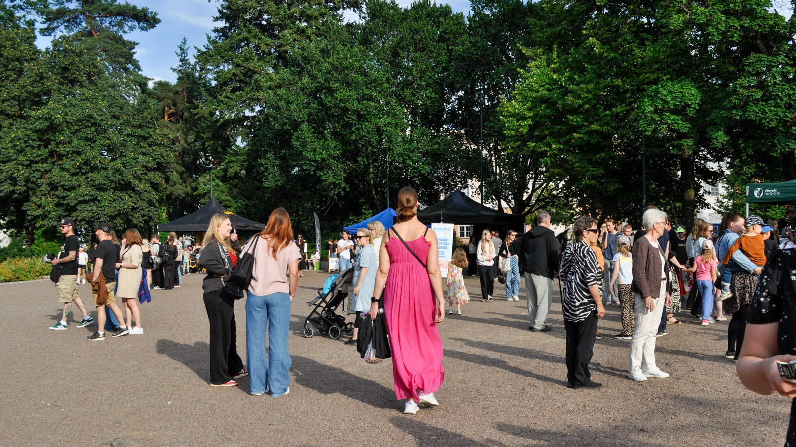 Ihmisiä Kouvolan Keskuspuistossa Taiteiden yössä. Kuvan keskellä on nainen vaaleanpunaisessa kesämekossa, taustalla on puita ja esittelypisteitä.